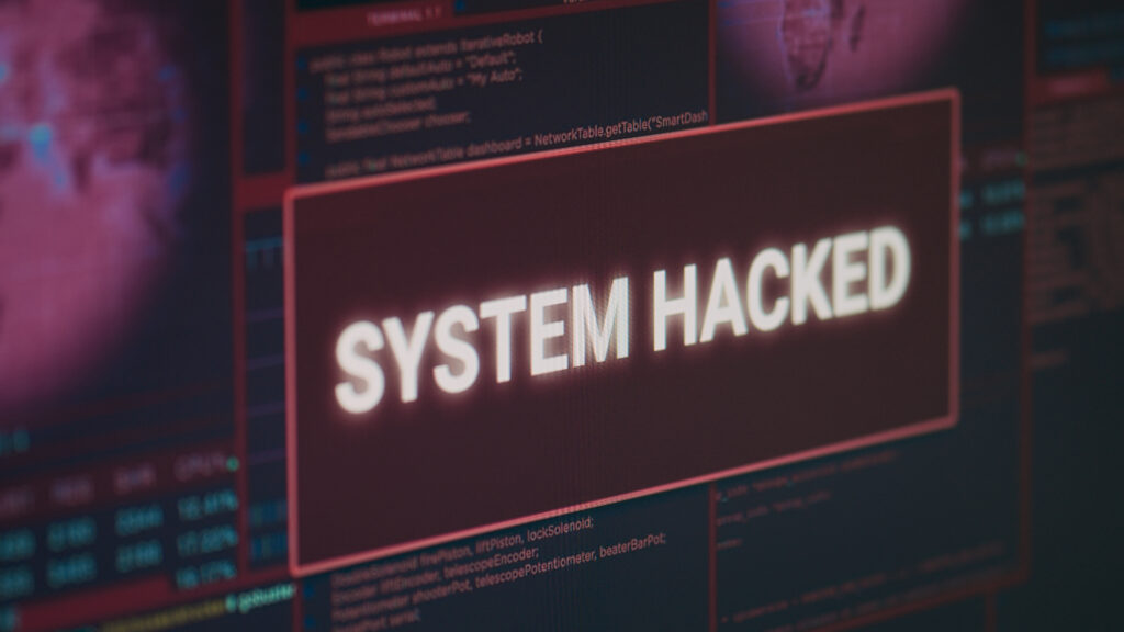 Auf dem Monitor erscheint „SYSTEM HACKED“ als Sicherheitswarnung, umgeben von rotem, leuchtendem Cyber-Alarm und Hinweisen zu Malware und Phishing.