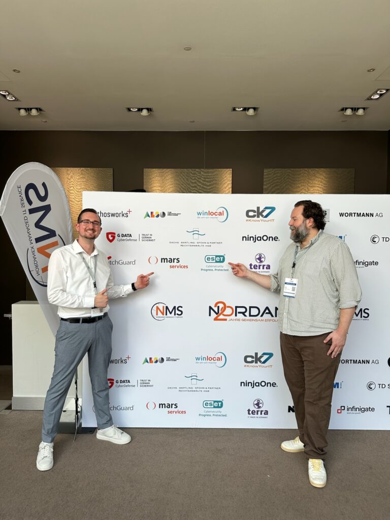 Zwei Teilnehmer des NMS Symposiums 2026 tauschen sich vor einem Marken-Backdrop mit mars services, NMS und weiteren IT-Partnern aus.