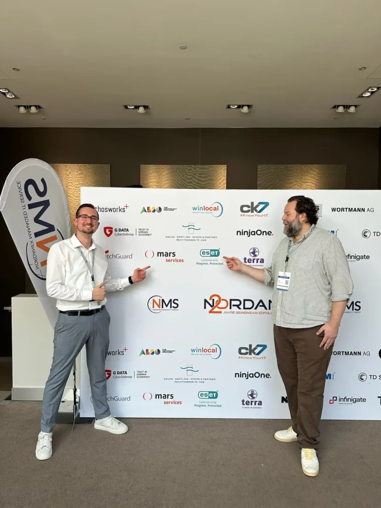 Zwei Männer zeigen lachend auf die Partnerlogo-Wand beim NMS Symposium zum 20-jährigen NORDANEX-Jubiläum